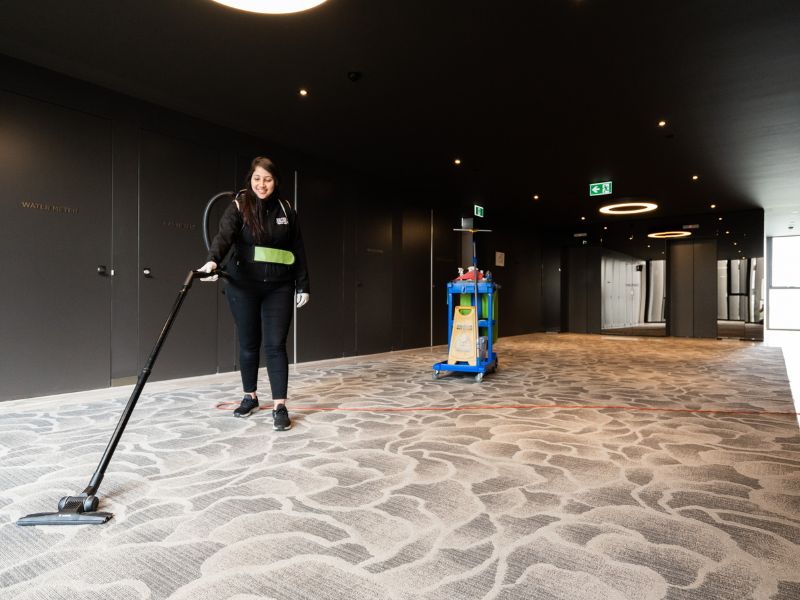 Smiling Commercial Cleaner Vaccumming Carpet In Communal Hallway Space - Commercial Cleaning South Yarra